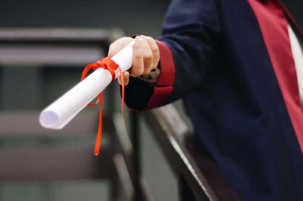  A graduate in a gown holding out a rolled diploma tied with a red ribbon, symbolizing educational success and the perfect occasion for giving meaningful investment gifts to support future aspirations.