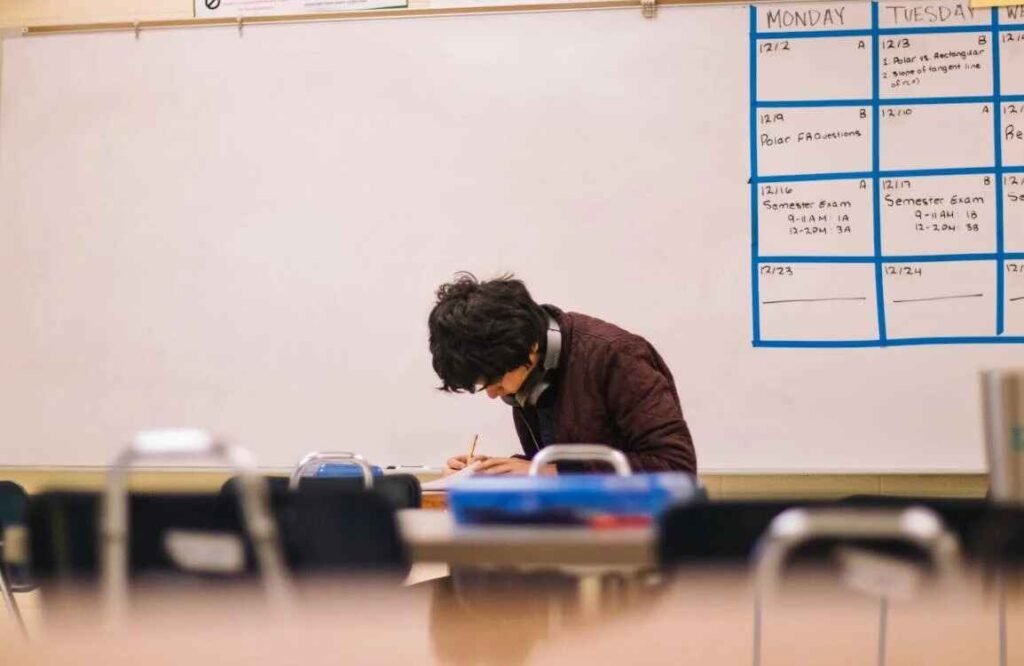 A high school student sitting alone in a quiet classroom, focused on writing in his notebook while wearing headphones around his neck. The classroom is empty, and a weekly schedule is visible on the whiteboard behind him. This image represents budgeting scenarios for high school students, symbolizing a student skipping a party or social event to prioritize academics and personal goals.