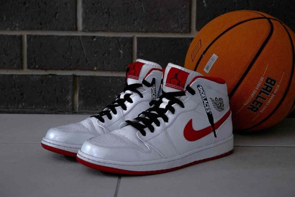A stylish pair of white and red Nike Air Jordan sneakers placed on a tiled floor next to an orange basketball with the word “BALLER” printed on it. This image is ideal for content related to illustrating the challenge of choosing between trendy, high-cost items like branded sneakers and managing a smart spending plan.