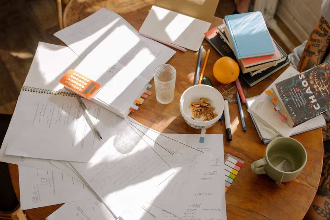 Some papers, a calculator, a bowl having walnuts, a pile of books, and some pencils are on a table representing budgeting activities for high school students.