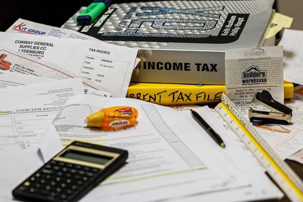 "A cluttered desk with tax invoices, receipts, and financial documents. In the foreground, a calculator and a pen rest on tax-related papers, while a 'CURRENT TAX FILES' folder sits next to them. This image emphasizes the importance of organizing financial information, which can be supported by using the best budgeting apps for teens to track and manage personal finances efficiently."
