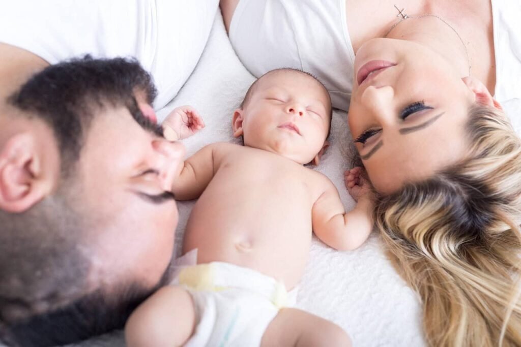"A peaceful moment of a young family lying on a white blanket, with a baby resting in the center between the father and mother. The baby is peacefully sleeping, and both parents are gazing lovingly at their child, creating a warm, intimate family scene, symbolizing the benefits of applying budgeting tips for young families.