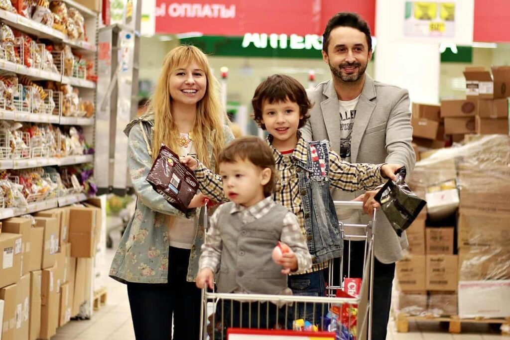 "A cheerful family of four shopping together in a store aisle. The mother, smiling and holding a pack of snacks, stands beside her two children, one of whom is holding a toy, while the father walks next to them, pushing the shopping cart. The background shows shelves filled with items and boxes, capturing a joyful family outing, emphasizing the benefits of adapting budgeting tips for young families.