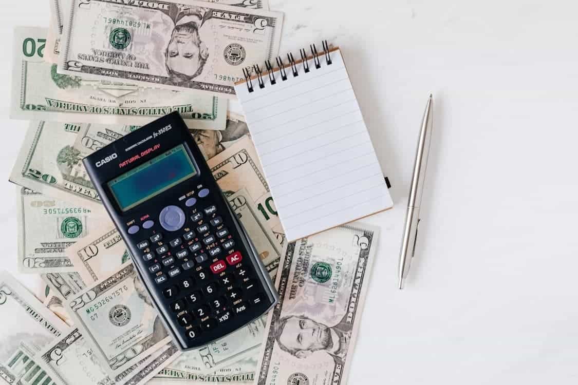 A financial calculator, a blank notepad, a pen, and various U.S. dollar bills are arranged on a white surface—symbolizing the tools students can use to budget effectively. This setup reflects active money management, planning, and tracking expenses, which are essential habits for students learning to take control of their finances.