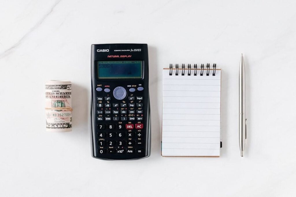 Flat lay of essential budgeting tools including a roll of cash, a scientific calculator, a notepad, and a pen on a white background — symbolizing the importance of using simple tools to track expenses, plan finances, and stay organized as a student.