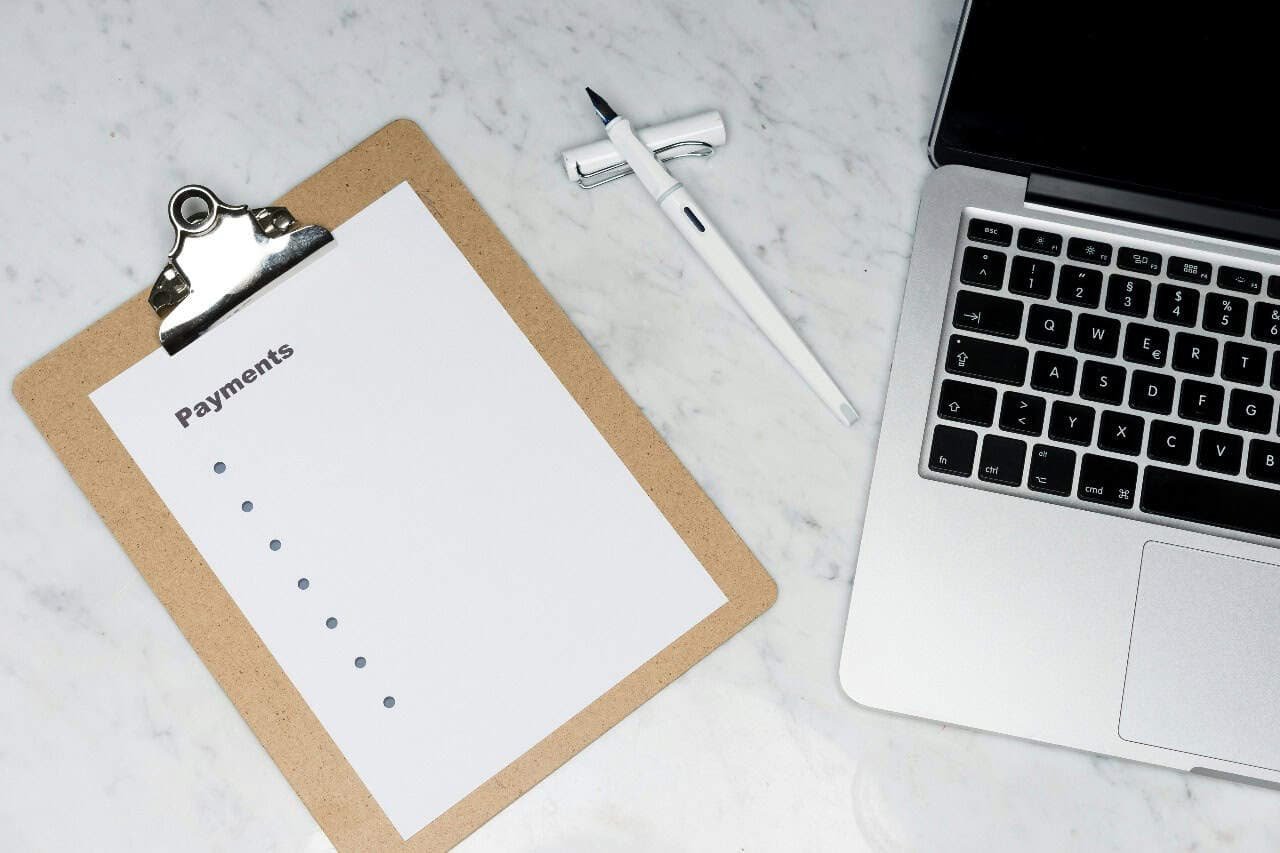 Budgeting setup on a marble desk featuring a clipboard with a 'Payments' checklist, a pen, and a laptop—symbolizing financial planning for life after high school."