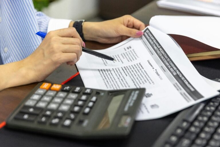 A person is using a pen to review financial documents while sitting at a desk. A calculator is placed nearby, emphasizing the importance of budgeting for life after high school."