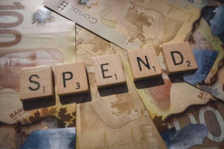Close-up image of wooden Scrabble tiles spelling “SPEND” placed on top of scattered Canadian 100-dollar bills, symbolizing personal finance and budgeting decisions. This visual evokes questions around money habits, making it ideal for content exploring how much spending a week is reasonable in a modern economic context.