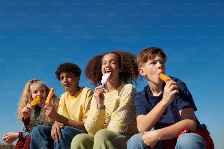 A group of four diverse teenagers sitting outdoors under a clear blue sky, enjoying colorful ice creams and laughing together, capturing a carefree moment of youthful fun. This relaxed scene prompts the question: how much spending a week is reasonable?—highlighting the balance between enjoying small treats and managing a weekly budget wisely.