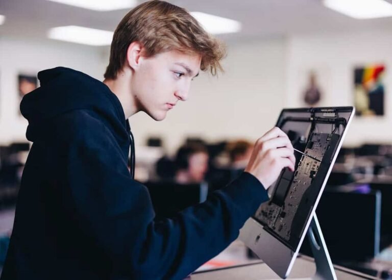 A focused teenage boy in a dark hoodie using a tool to repair or examine the internal components of a desktop computer in a classroom or tech lab setting.