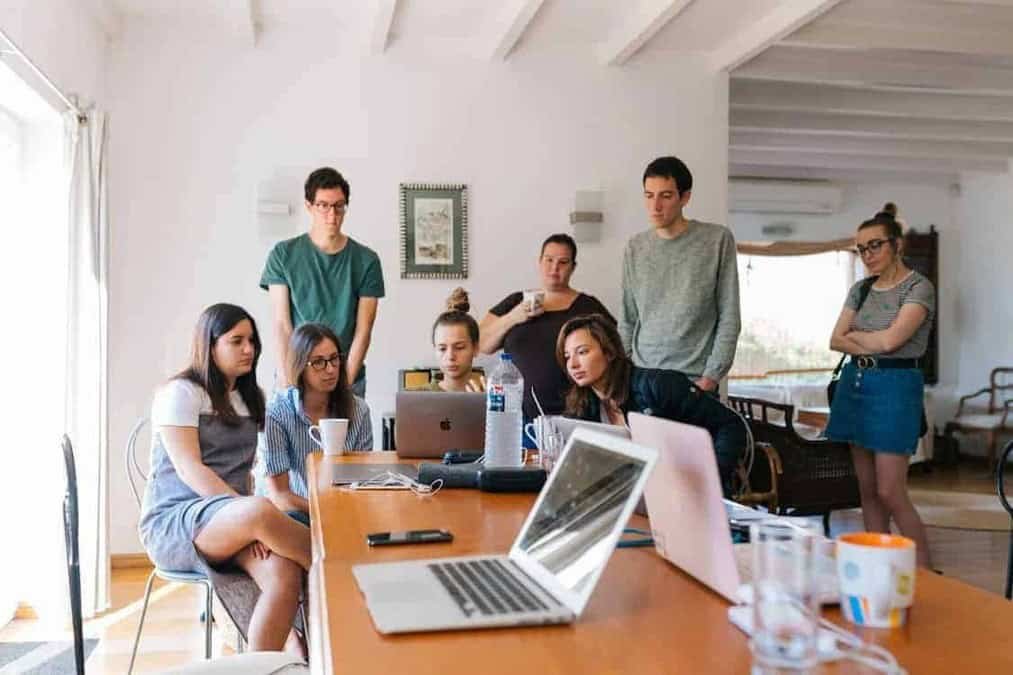 A diverse group of young adults, including both men and women, are gathered around a wooden table in a well-lit room, intently focused on laptops and a tablet. The image captures a collaborative session, with some individuals seated and others standing, suggesting a dynamic and engaged discussion. The scene could be interpreted as a study group, a startup meeting, or a financial planning workshop. The image's content and context make it an ideal visual aid for articles and resources about youth financial literacy, collaborative work, and modern education.
