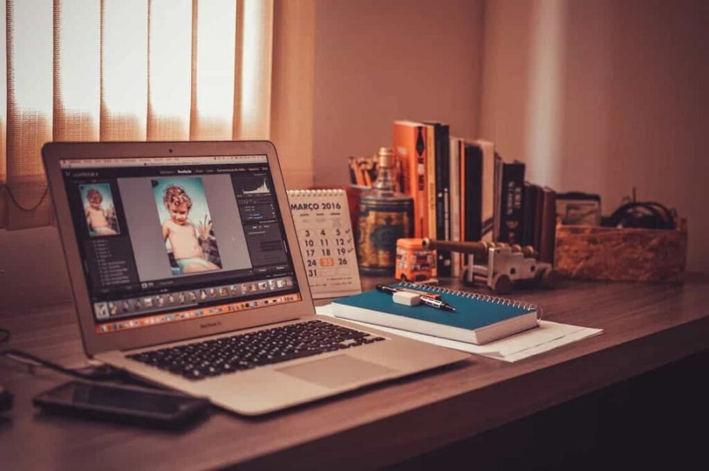 A MacBook laptop is open on a desk, displaying an image of a young child on the screen, likely in a photo editing program. A calendar for March 2016, a stack of books, and various other desk items, including a notepad and a pen, are visible next to the laptop.