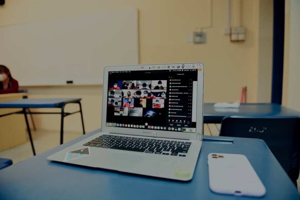 A classroom setting with a laptop and a smartphone on a desk. The laptop screen displays a video conference call, likely a virtual classroom or meeting, with multiple participants visible in small squares. The setting suggests a hybrid learning or work environment, where a physical classroom is being used for an online session. This image could be used to illustrate topics related to education technology, remote learning, hybrid classrooms, or the integration of digital tools in traditional learning spaces.