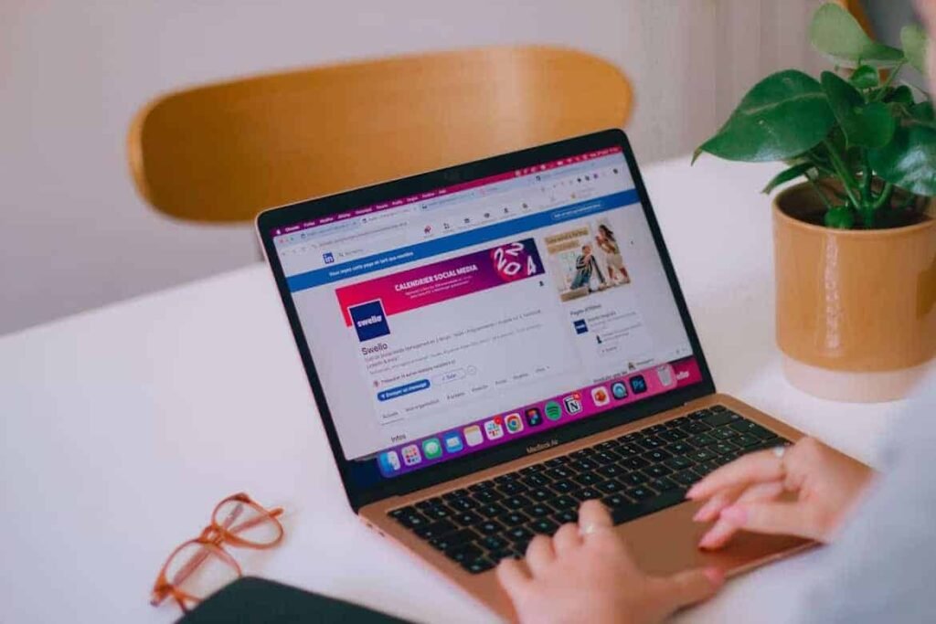 Close-up of a person using a MacBook Air to browse LinkedIn, with the screen displaying a company page for "Swello" focused on social media content. The workspace includes a potted green plant, eyeglasses, and a notebook on a white desk. This image illustrates a modern digital work environment, ideal for remote opportunities and online jobs for teens interested in social media or digital marketing. 