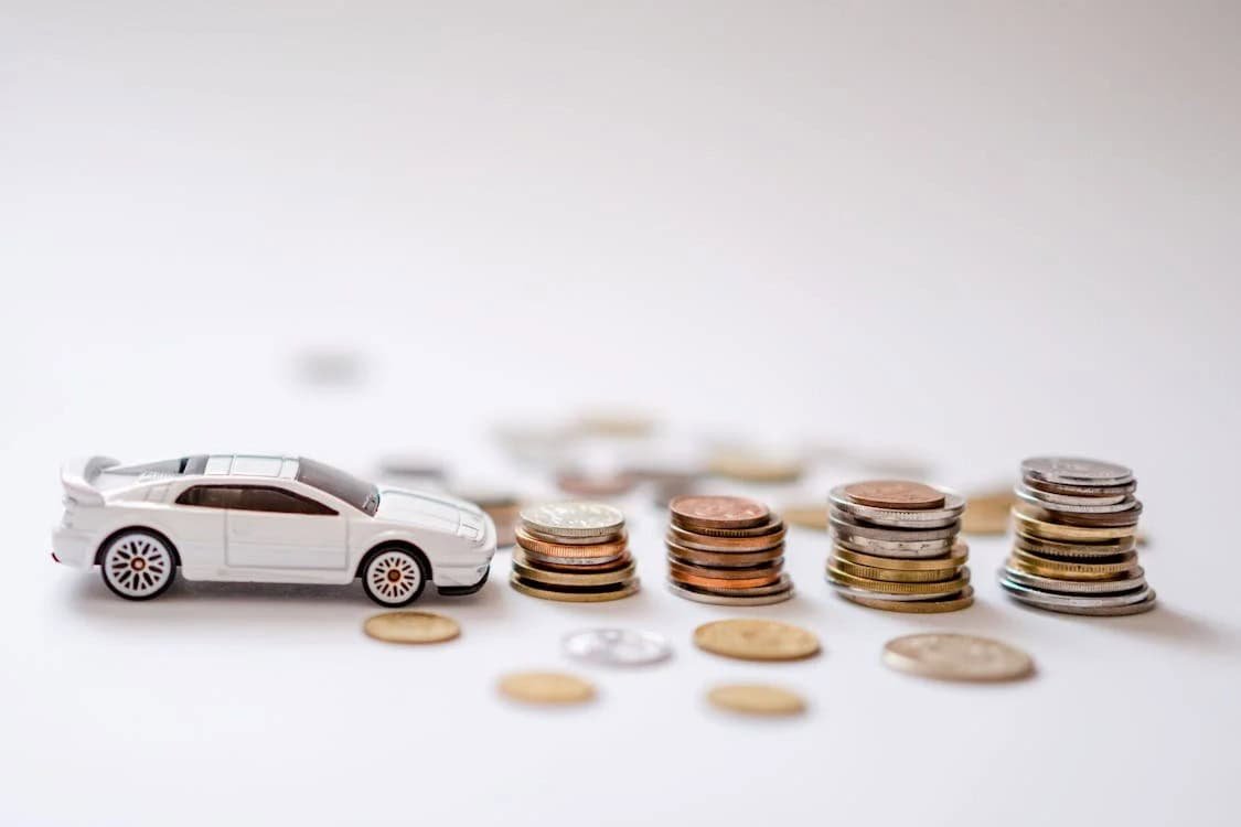 A white toy car beside ascending stacks of coins on a white background, symbolizing saving money for a car.