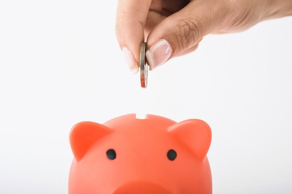 A close-up of a hand dropping a coin into an orange piggy bank, symbolizing saving money and building financial habits as a teenager.