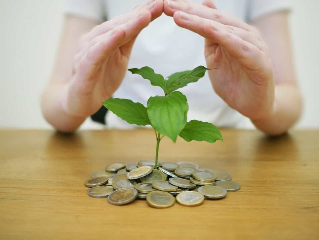 Hands form a protective shelter over a small green plant growing from a pile of coins on a wooden table. This image represents the concept of nurturing and growing savings, and could be used for content related to personal finance for teens, smart spending, and building wealth from a young age.