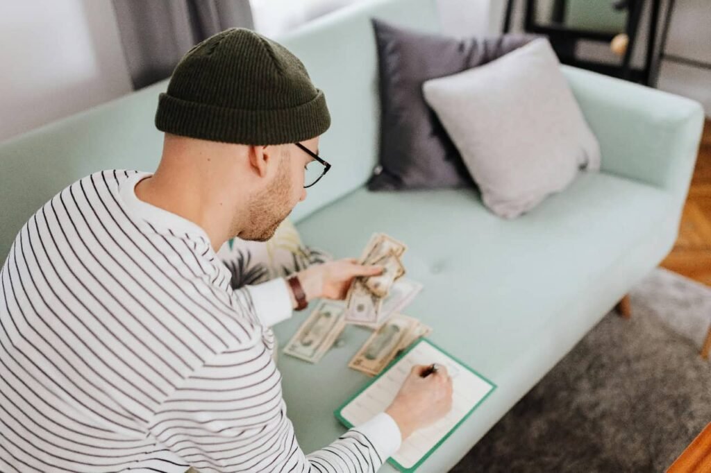 A man wearing a green beanie and glasses sits on the floor in front of a light green couch. He is holding several dollar bills in one hand and is using the other hand to write in a small green notebook. Other stacks of money are laid out on the floor in front of him. The image visually represents the concept of budgeting and managing personal finances.A man wearing a green beanie and glasses sits on the floor in front of a light green couch. He is holding several dollar bills in one hand and is using the other hand to write in a small green notebook. Other stacks of money are laid out on the floor in front of him. The image visually represents the concept of budgeting and managing personal finances.A man wearing a green beanie and glasses sits on the floor in front of a light green couch. He is holding several dollar bills in one hand and is using the other hand to write in a small green notebook. Other stacks of money are laid out on the floor in front of him. The image visually represents the concept of budgeting and managing personal finances.A man wearing a green beanie and glasses sits on the floor in front of a light green couch. He is holding several dollar bills in one hand and is using the other hand to write in a small green notebook. Other stacks of money are laid out on the floor in front of him. The image visually represents the concept of budgeting and managing personal finances.A man wearing a green beanie and glasses sits on the floor in front of a light green couch. He is holding several dollar bills in one hand and is using the other hand to write in a small green notebook. Other stacks of money are laid out on the floor in front of him. The image visually represents the concept of budgeting and managing personal finances.A man wearing a green beanie and glasses sits on the floor in front of a light green couch. He is holding several dollar bills in one hand and is using the other hand to write in a small green notebook. Other stacks of money are laid out on the floor in front of him. The image visually represents the concept of budgeting and managing personal finances.A man wearing a green beanie and glasses sits on the floor in front of a light green couch. He is holding several dollar bills in one hand and is using the other hand to write in a small green notebook. Other stacks of money are laid out on the floor in front of him. The image visually represents the concept of budgeting and managing personal finances.
