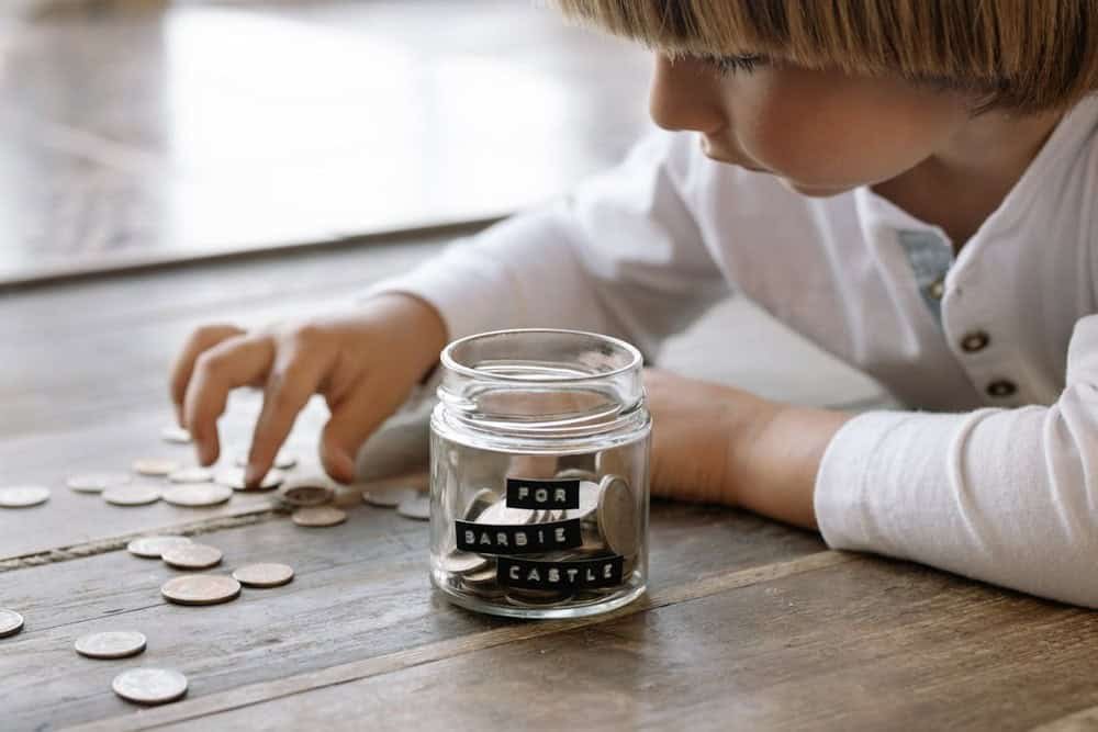 A young teen carefully counts coins beside a glass savings jar labeled “For Barbie Castle,” symbolizing early saving habits and financial planning for personal goals.