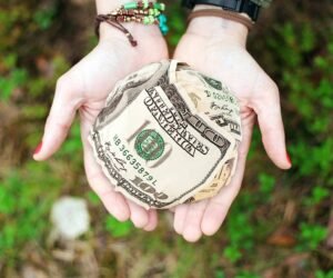 A person’s hands are cupped together, holding a large, crumpled ball of U.S. dollar bills. This image represents the importance of managing cash wisely, an essential skill for teens learning to budget.