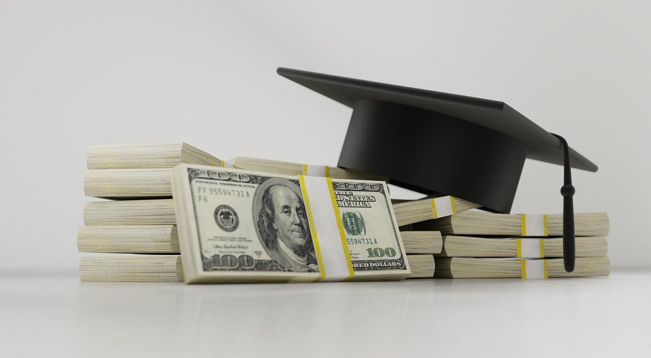 Graduation cap on stacks of $100 bills symbolizing student debt and college costs