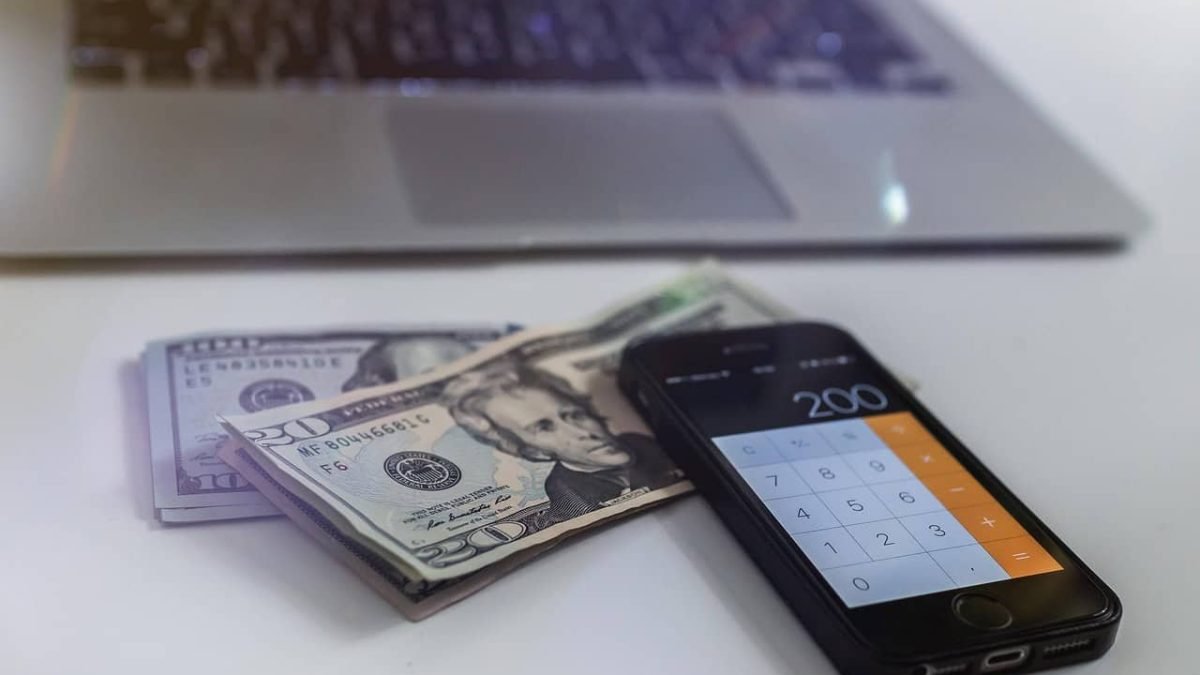 The image features a close-up shot of a smartphone displaying a calculator app with the value "200" visible on the screen. Next to the phone, there are several bills, including a $100 bill and a $20 bill, placed on a desk. In the background, a laptop is partially visible. The image is focused on the items on the desk, with a soft blur effect on the laptop, showing the importance of regular checking and modifying the budget to avoid common budgeting mistakes.